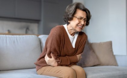 an older woman sits on a sofa clutching her stomach